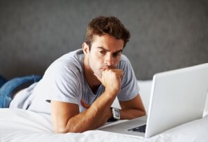 Portrait of a thoughtful young man using laptop while lying on bed at home - Indoor
